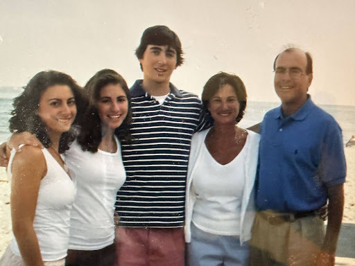 Five smiling family members stand together on a beach. Two young women in white shirts, a young man in a striped polo and pink shorts, an older woman in a white top, and an older man in a blue polo and khakis. The ocean is in the background.