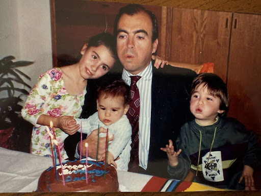 Father in suit blows out lit candles on a chocolate birthday cake with "Happy Birthday" text. A smiling girl in a floral top leans on him, a baby watches the cake, and a boy in a green "Geographic World" sweater observes with raised hand.