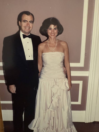 A smiling man in a black tuxedo and bow tie stands with his arm around a smiling woman in a strapless light pink ruffled gown and pearl necklace.