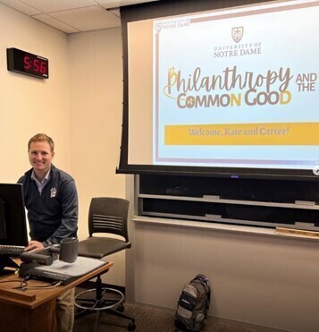 Smiling man in blue Notre Dame polo sits at desk. Screen: Philanthropy and the Common Good, Welcome Kate and Carter!