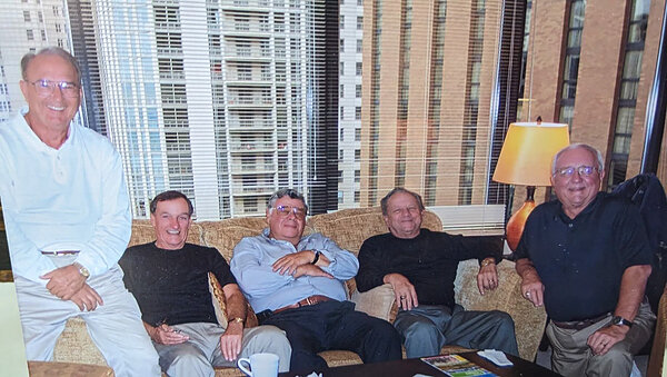 Five men in casual attire sit on a sofa in front of a window with blinds and city buildings.